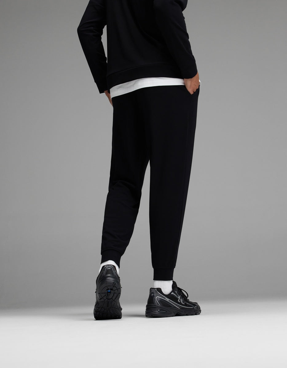 A person stands facing away, wearing black jogger pants, a black jacket, a white shirt, white socks, and black sneakers against a gray background.|Color:Black