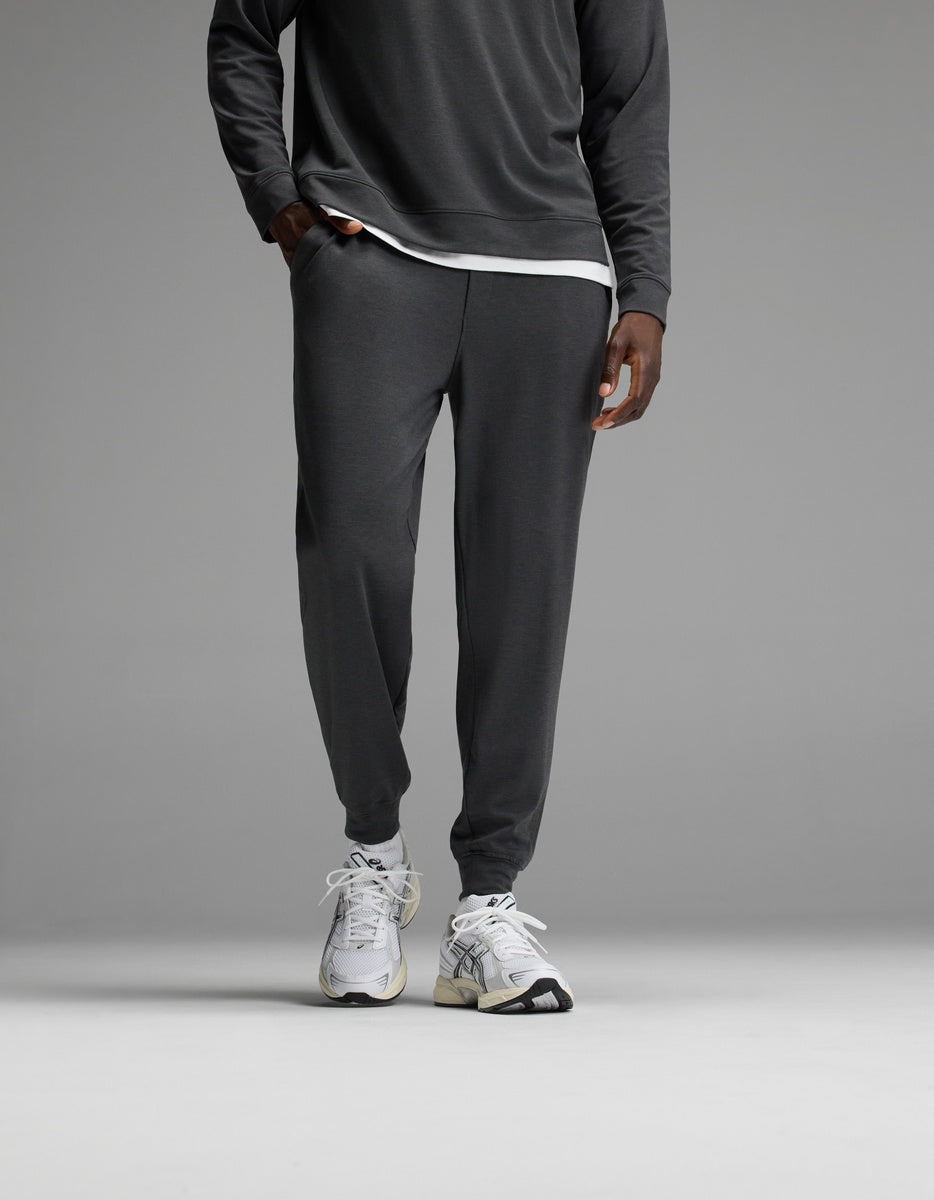 A person stands against a plain gray background wearing Cozy Earth’s Men’s Brushed Bamboo Joggers in Charcoal, paired with a matching long-sleeve top layered over a white undershirt and finished with white athletic sneakers. The upper body is partially out of frame.|Color:Charcoal