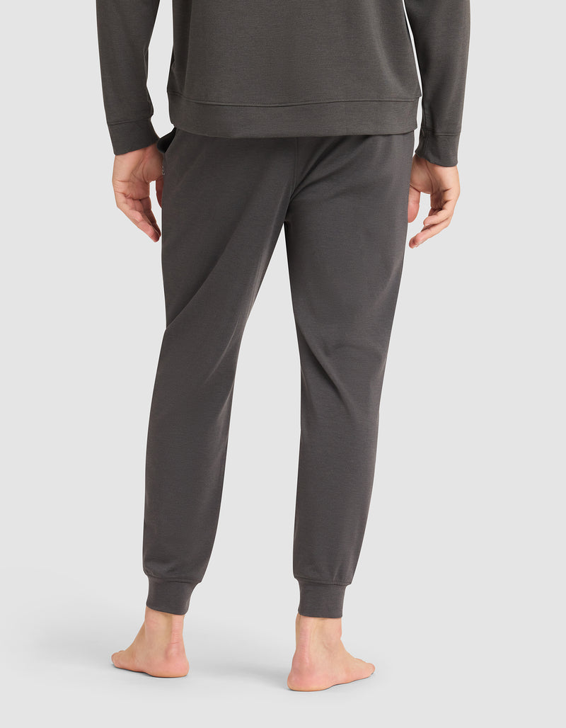 A person stands barefoot on a white background, shown from the back wearing Cozy Earth's Men's Brushed Bamboo Jogger Pant in dark gray paired with a matching long-sleeve top.