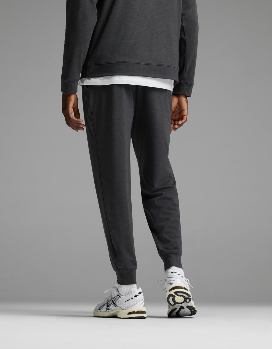 A person stands facing away, wearing the Cozy Earth Men’s Brushed Bamboo Joggers and a matching dark sweatshirt, paired with white sneakers featuring black details, against a plain gray background.