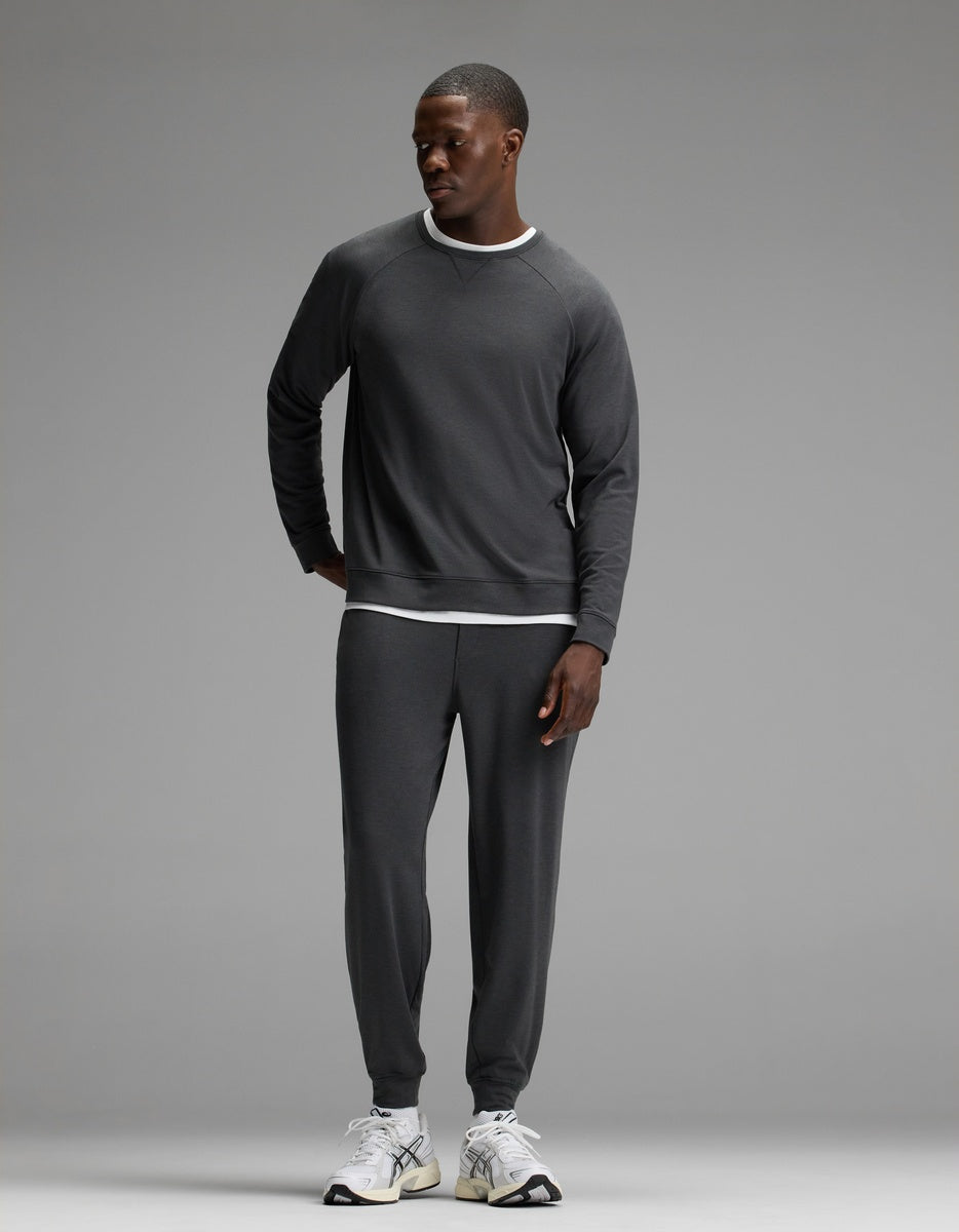 A man stands against a plain gray background, wearing Cozy Earth’s Men’s Brushed Bamboo Joggers in dark gray with a matching sweatshirt layered over a white T-shirt and paired with white sneakers. He looks to his left with one hand behind his back.