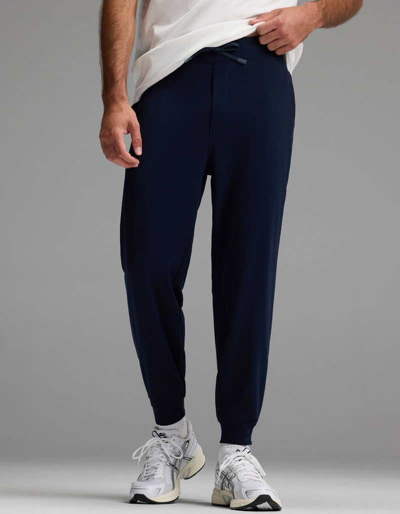 A person stands against a plain gray background, wearing Cozy Earth Men’s Brushed Bamboo Joggers in navy blue, along with a white T-shirt and white athletic sneakers. Only the lower half of the body is visible.