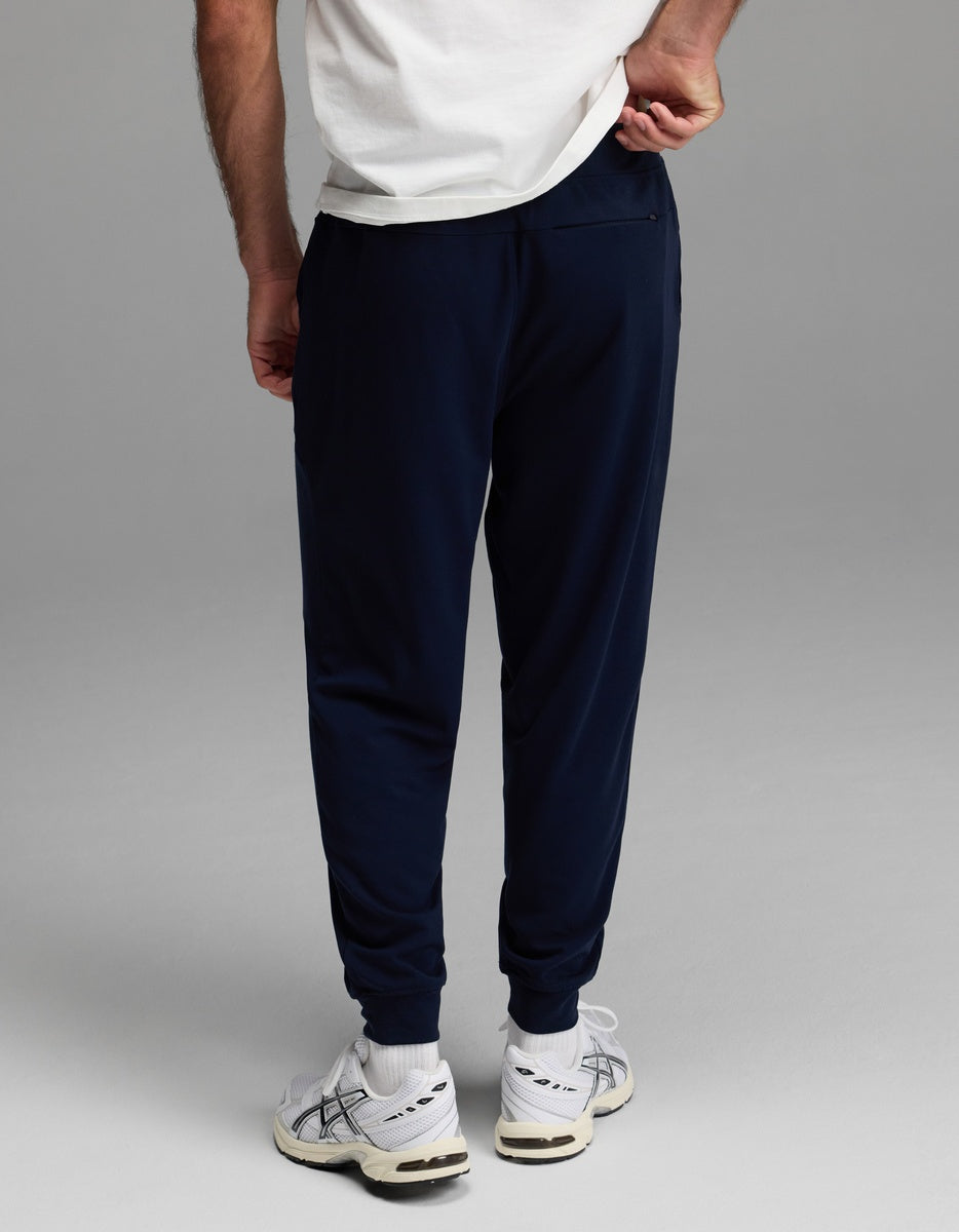 A person stands facing away, wearing a white T-shirt, white socks, white sneakers, and Cozy Earth’s Men’s Brushed Bamboo Joggers in navy, with one hand pulling at the back of their shirt.
