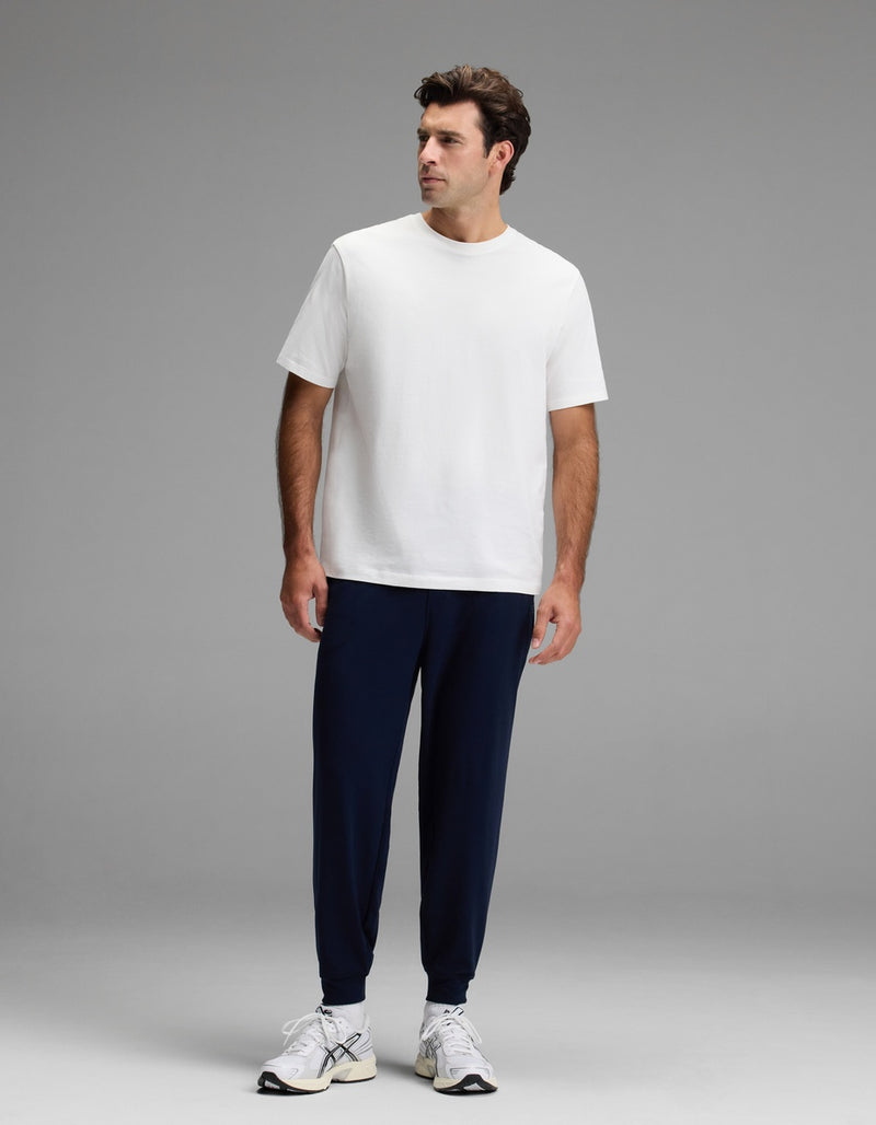 A man stands against a grey background, wearing a plain white t-shirt, white sneakers, and Cozy Earth’s Men’s Brushed Bamboo Joggers in dark blue. He gazes slightly to his left with a neutral expression.