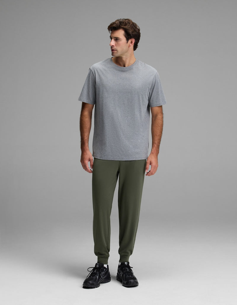 A man stands in a studio against a neutral grey background, looking off to his left while wearing Cozy Earth’s Men’s Brushed Bamboo Joggers in olive green, paired with a plain grey t-shirt and black sneakers.