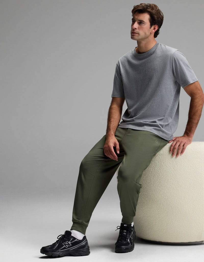 A man in a gray t-shirt and black sneakers sits on a round, white textured ottoman against a plain gray background, wearing Cozy Earth Men’s Brushed Bamboo Joggers in olive.