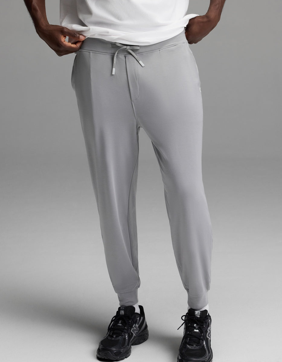 A person is shown from the shoulders down, wearing Cozy Earth’s Men’s Brushed Bamboo Joggers in light gray, paired with a white shirt and black athletic shoes, standing against a plain gray background.|Color:Stone