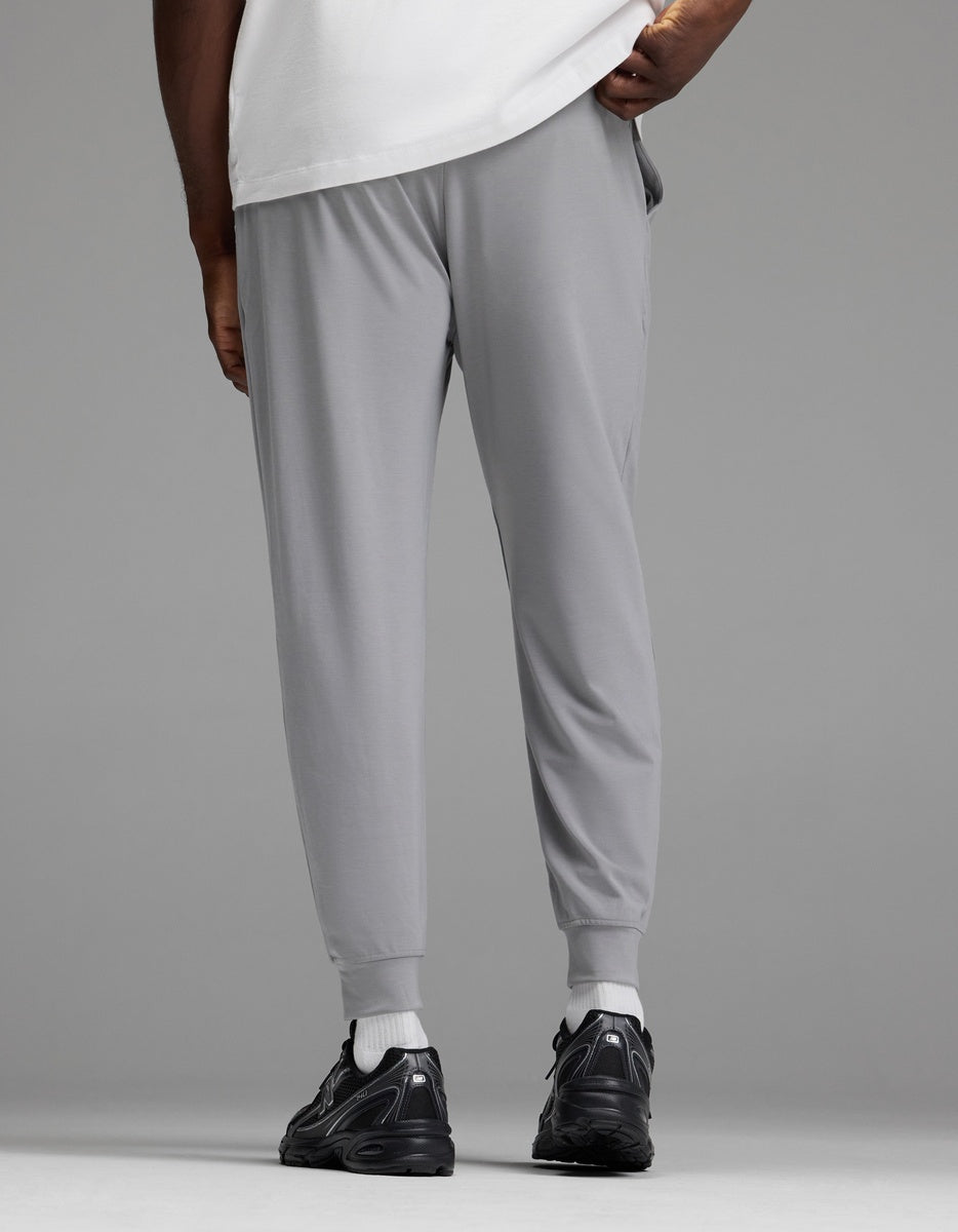 A person is shown from the back, standing against a plain gray background, wearing Cozy Earth Men’s Brushed Bamboo Joggers in light gray paired with a white shirt and black sneakers.|Color:Stone