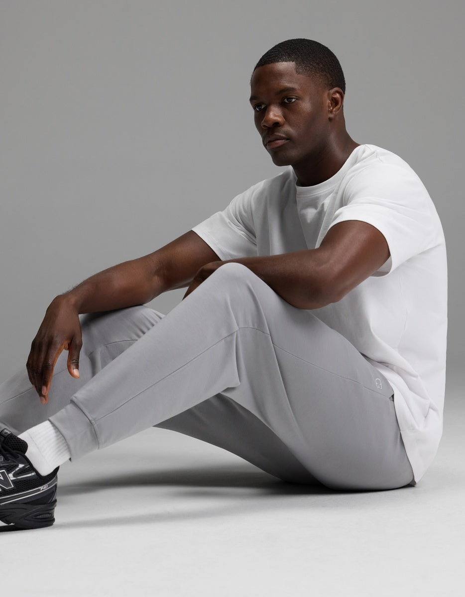 A man wearing Cozy Earth’s Men’s Brushed Bamboo Joggers in light gray, paired with a white t-shirt, white socks, and black sneakers, sits on the floor against a plain gray background.