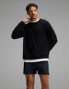 A man stands against a gray background, wearing the Cozy Earth Men’s Brushed Bamboo Pullover Crew in black over a white shirt and black shorts. He has short curly hair and is touching his neck with his right hand, looking slightly to the side.|Color:Black