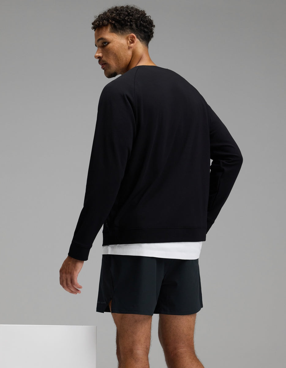 A man with short curly hair is wearing the Cozy Earth Men’s Brushed Bamboo Pullover Crew in black over a white shirt and paired with black shorts, standing and looking over his shoulder against a plain gray background.|Color:Black