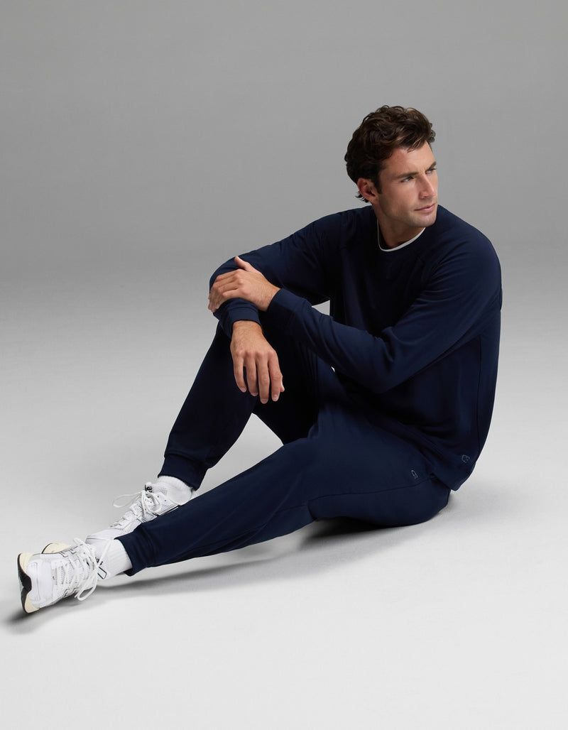 A man wearing the Cozy Earth Men’s Brushed Bamboo Jogger Set in navy blue and white sneakers sits on the floor with one leg bent and the other extended, looking off to the side against a plain gray background.