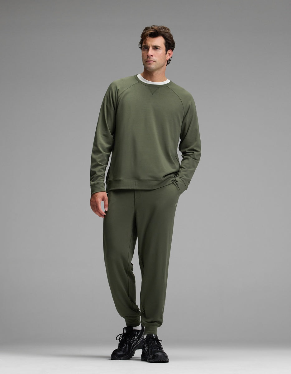 A man wearing the Cozy Earth Men’s Brushed Bamboo Jogger Set in olive green, paired with black sneakers, stands against a plain gray background, posing with one hand in his pocket and gazing slightly to the side.|Color:Olive
