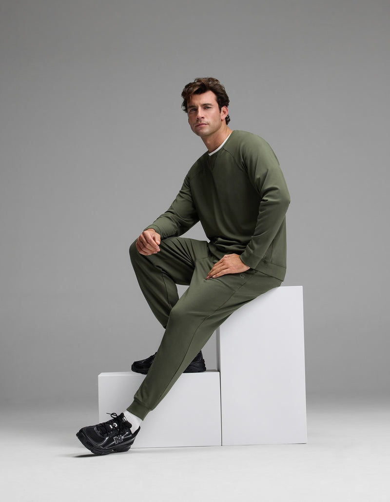 A man wearing the Cozy Earth Men’s Brushed Bamboo Jogger Set in olive green and black sneakers sits on white geometric blocks against a plain gray background, gazing slightly to the side.