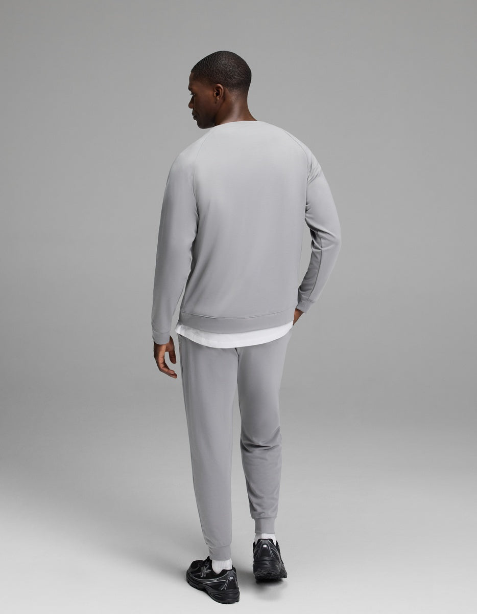 A man stands facing away, wearing the Cozy Earth Men’s Brushed Bamboo Jogger Set in light gray paired with black sneakers, his left hand resting on his hip against a plain, neutral gray background.|Color:Stone