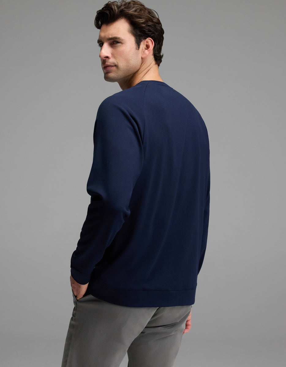 A man with dark hair, dressed in the Cozy Earth Men’s Brushed Bamboo Pullover Crew in navy and gray pants, stands with his back to the camera and glances over his shoulder against a plain gray background.
