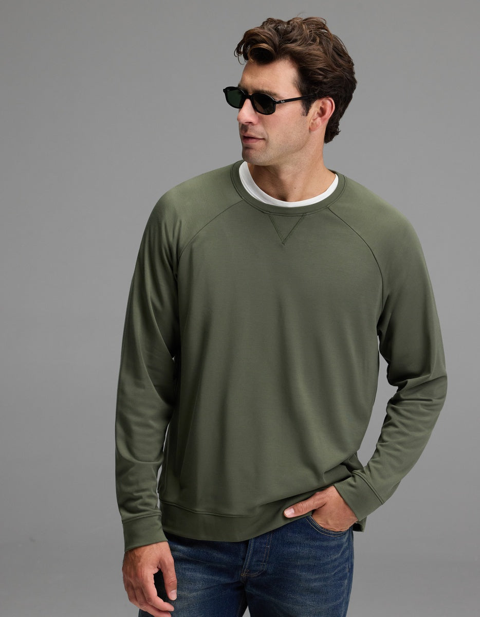 A man stands against a plain gray background with one hand in his jeans pocket, wearing dark sunglasses, a white T-shirt, and the Cozy Earth Men’s Brushed Bamboo Pullover Crew in Olive.|Color:Olive