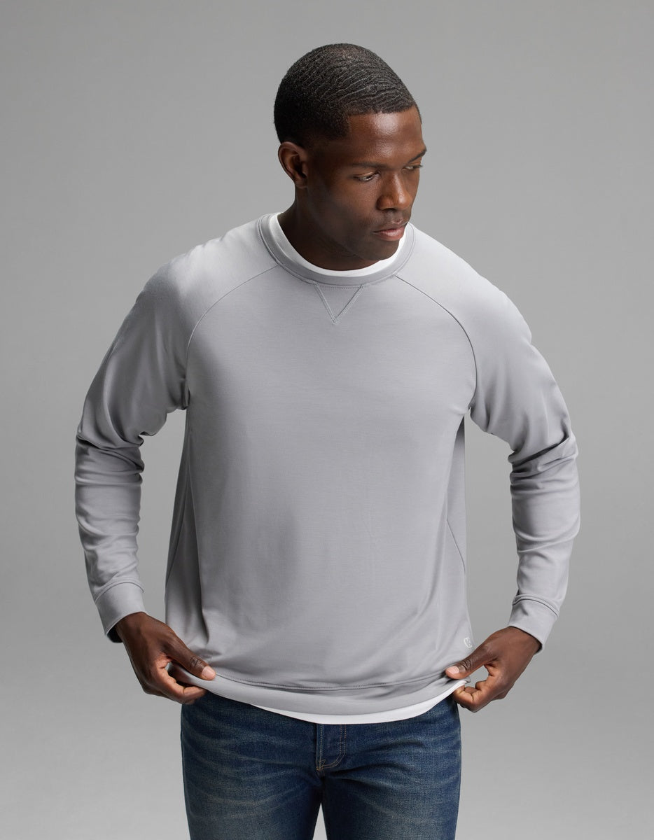 A man wearing the Cozy Earth Men’s Brushed Bamboo Pullover Crew in Stone adjusts his shirt while standing against a plain gray background.