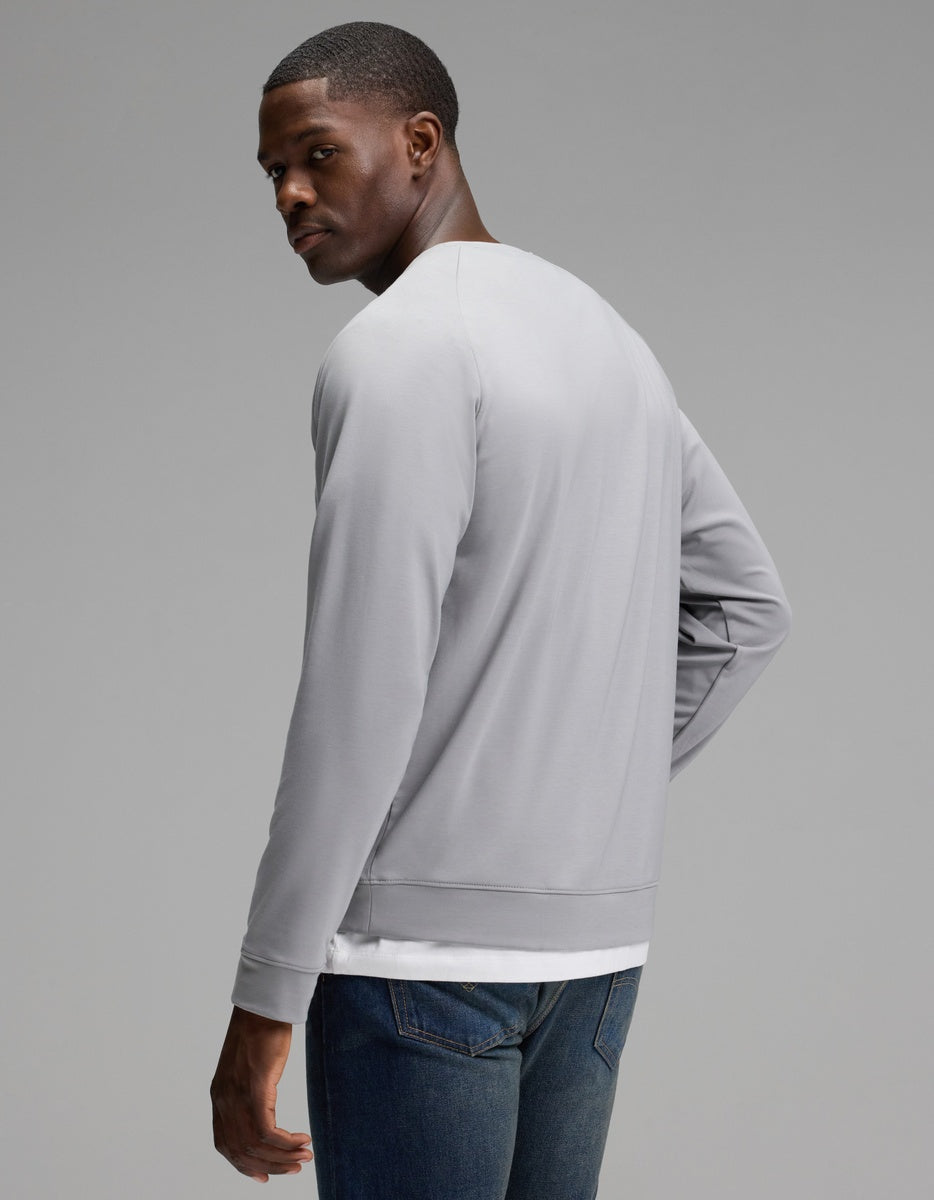 A man models the Cozy Earth Men’s Brushed Bamboo Pullover Crew in light gray, paired with blue jeans. He stands slightly turned away, glancing over his shoulder with a neutral expression against a plain gray background.