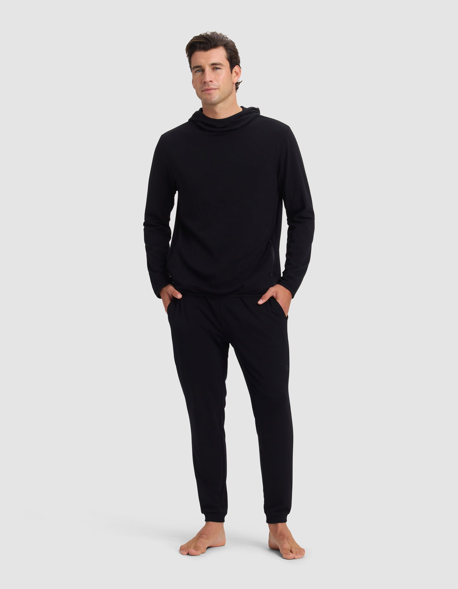 A man stands barefoot against a plain background, wearing the Cozy Earth Men's Brushed Bamboo Recovery Hoodie & Jogger Set in black. His hands are in his pockets and he gazes forward with a neutral expression.