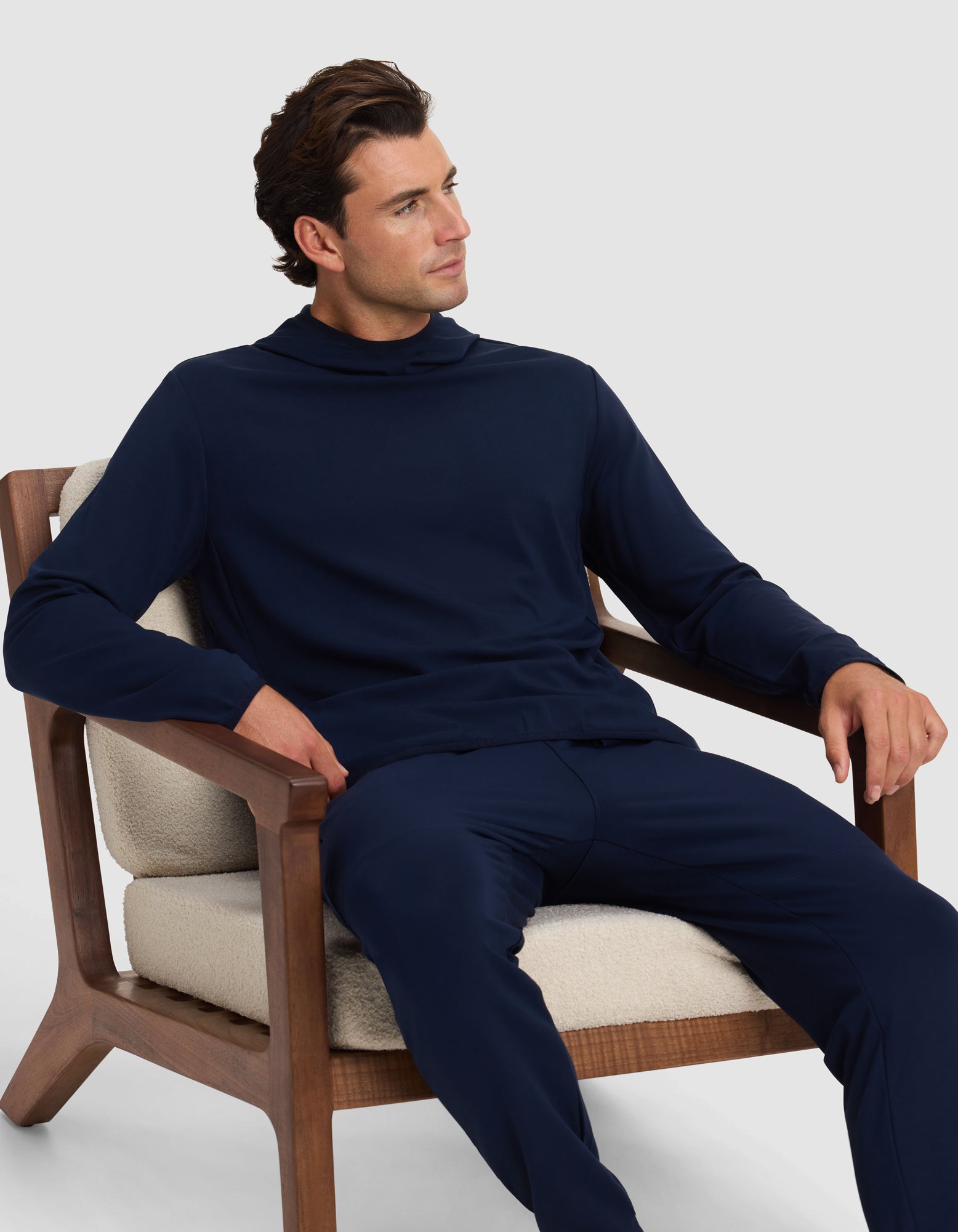 A man in a Cozy Earth Men's Brushed Bamboo Recovery Hoodie and matching pants sits on a cushioned wooden chair, looking left with a relaxed expression against a plain light background. 