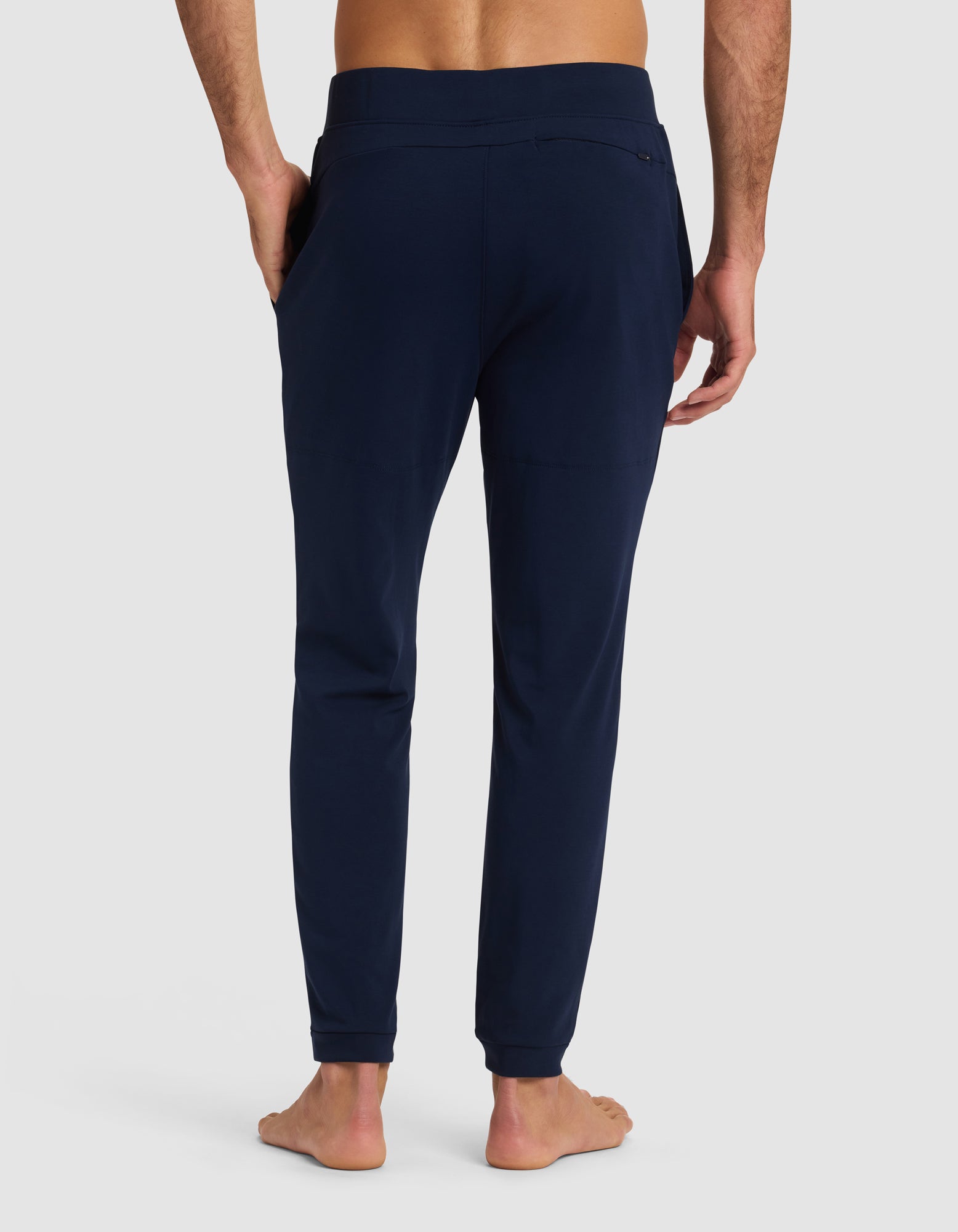 A barefoot man is shown from the back wearing Cozy Earth's Men's Brushed Bamboo Recovery Jogger in dark blue, with his hands in the side pockets, set against a plain light background.|Color:Navy