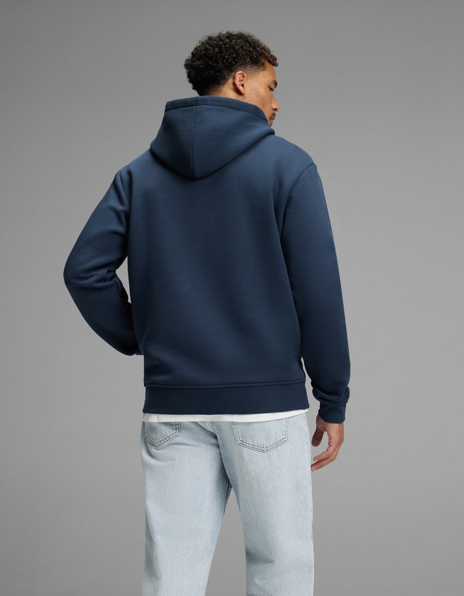 A person stands facing away, wearing the Cozy Earth Men’s Cityscape Hoodie in navy blue and light blue jeans, against a plain gray background.