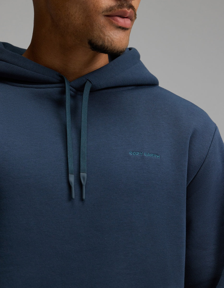 A person wearing the Men’s Cityscape Hoodie by Cozy Earth in dark blue, shown from the shoulders to the mouth, with “Cozy Earth” embroidered on the chest and hood strings hanging down.