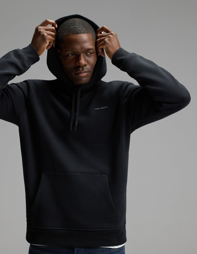 A man wears the Cozy Earth Men’s Cityscape Hoodie in Jet Black, lifting the hood over his head as he stands against a plain gray background, gazing to the side with a neutral expression.