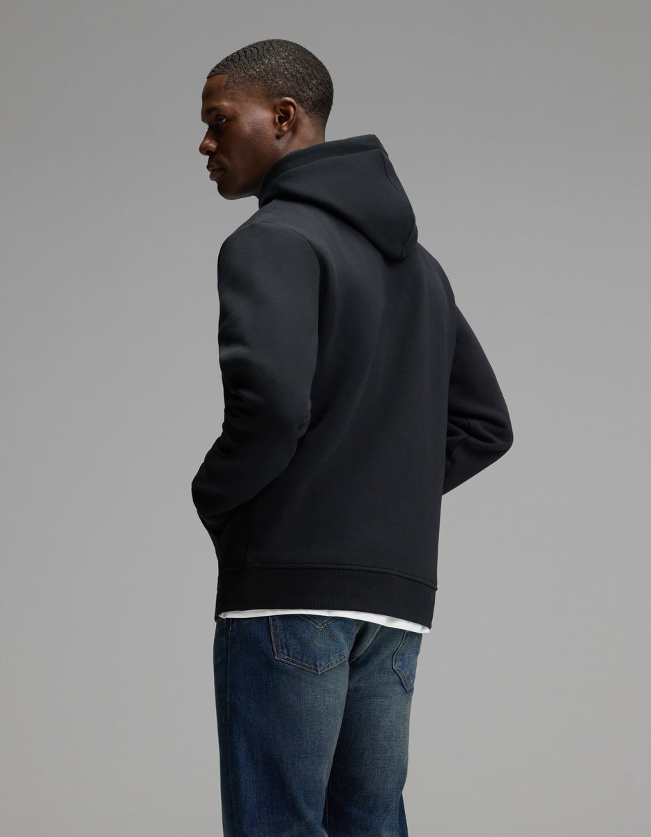 A person wearing the Cozy Earth Men’s Cityscape Hoodie in black and blue jeans stands with hands in pockets, facing away and glancing to the side against a plain gray background.