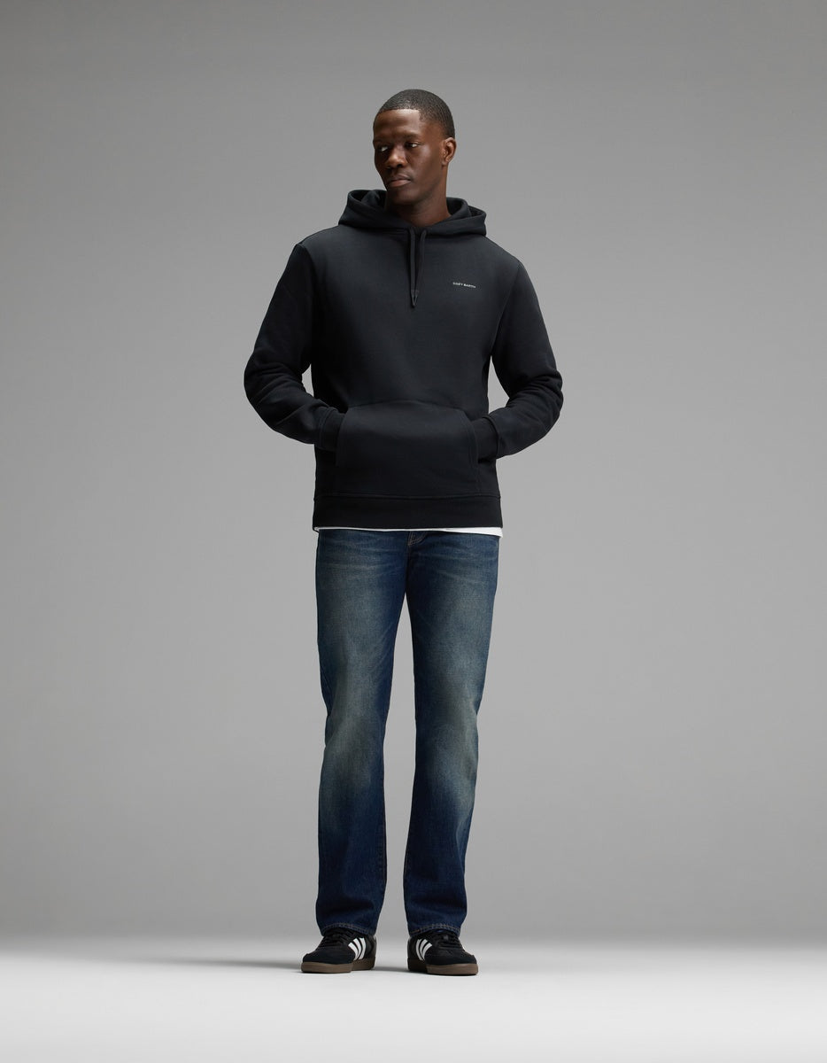 A man stands facing forward, wearing the Cozy Earth Men’s Cityscape Hoodie in black, paired with blue jeans, black slide sandals with white stripes, and a white shirt visible at the hem. The background is plain and gray.