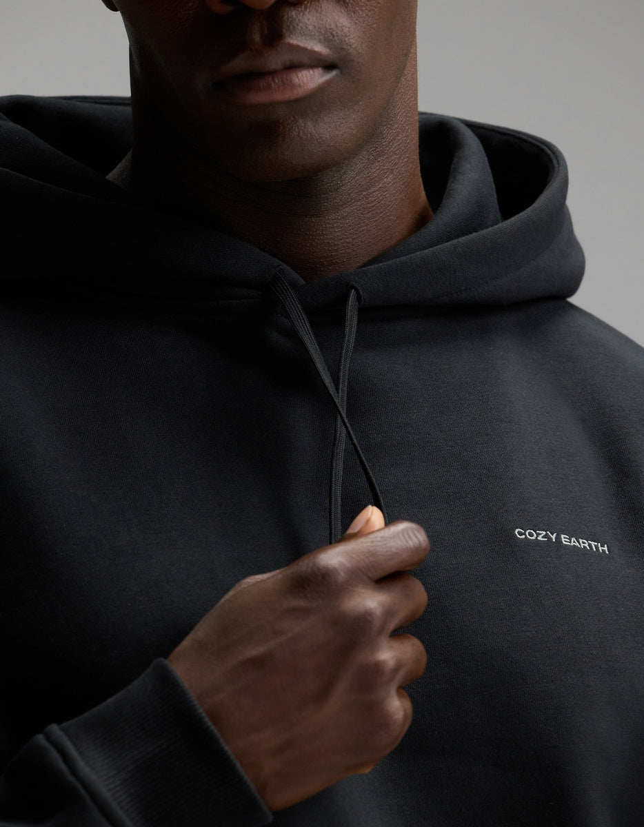 A person wearing the Cozy Earth Men’s Cityscape Hoodie in black, featuring the “COZY EARTH” logo, holds one of the drawstrings. The photo is cropped to show only the lower half of their face and upper torso.