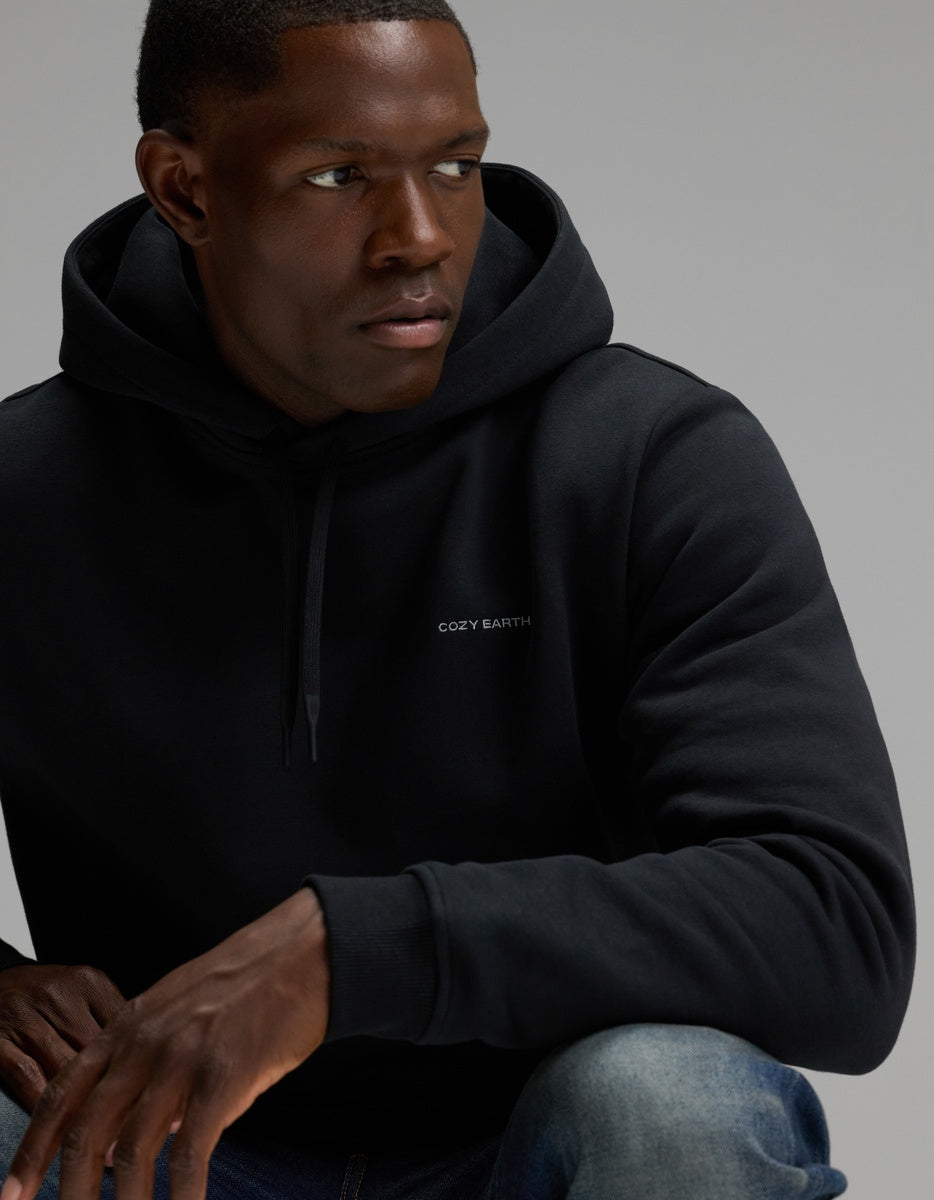 A man wearing the Men’s Cityscape Hoodie by Cozy Earth, featuring the brand’s text on the front, looks to his left with one arm resting on his knee against a plain background.