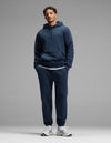 A person stands against a plain gray background, wearing the Cozy Earth Men’s Cityscape Hoodie & Sweatpant Set in Eclipse, paired with white sneakers and a white undershirt. Their hands are in their pockets and they are looking straight ahead.|Color:Eclipse