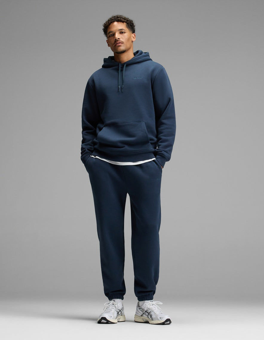 A person stands against a plain gray background, wearing the Cozy Earth Men’s Cityscape Hoodie & Sweatpant Set in Eclipse, paired with white sneakers and a white undershirt. Their hands are in their pockets and they are looking straight ahead.