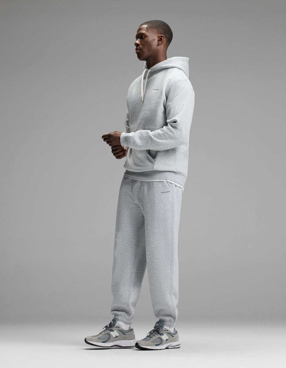 A person stands in profile against a gray background, wearing the Cozy Earth Men’s Cityscape Hoodie & Sweatpant Set paired with gray sneakers. The outfit looks casual and comfortable.|Color:Heather Grey