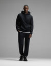 A man wearing Cozy Earth’s Men’s Cityscape Hoodie & Sweatpant Set in Jet Black stands against a plain gray background, facing the camera with a neutral expression.|Color:Jet Black