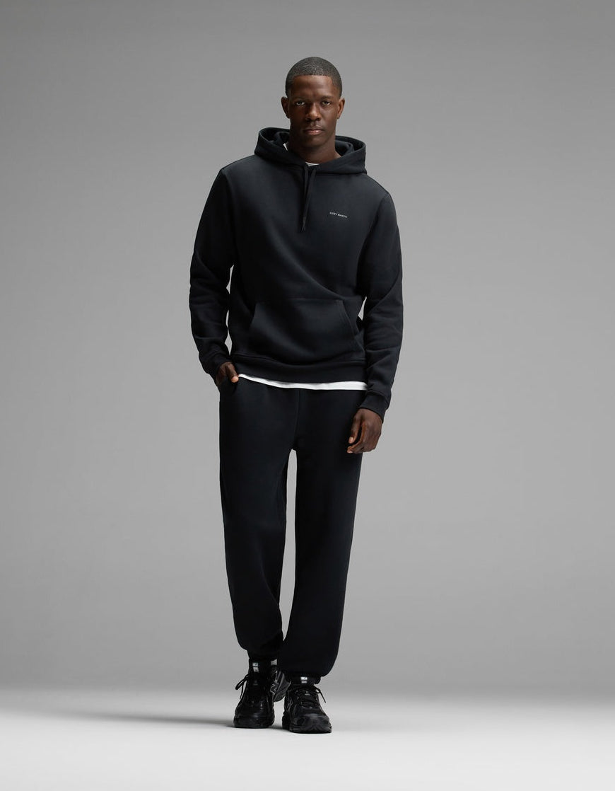A man wearing Cozy Earth’s Men’s Cityscape Hoodie & Sweatpant Set in Jet Black stands against a plain gray background, facing the camera with a neutral expression.|Color:Jet Black