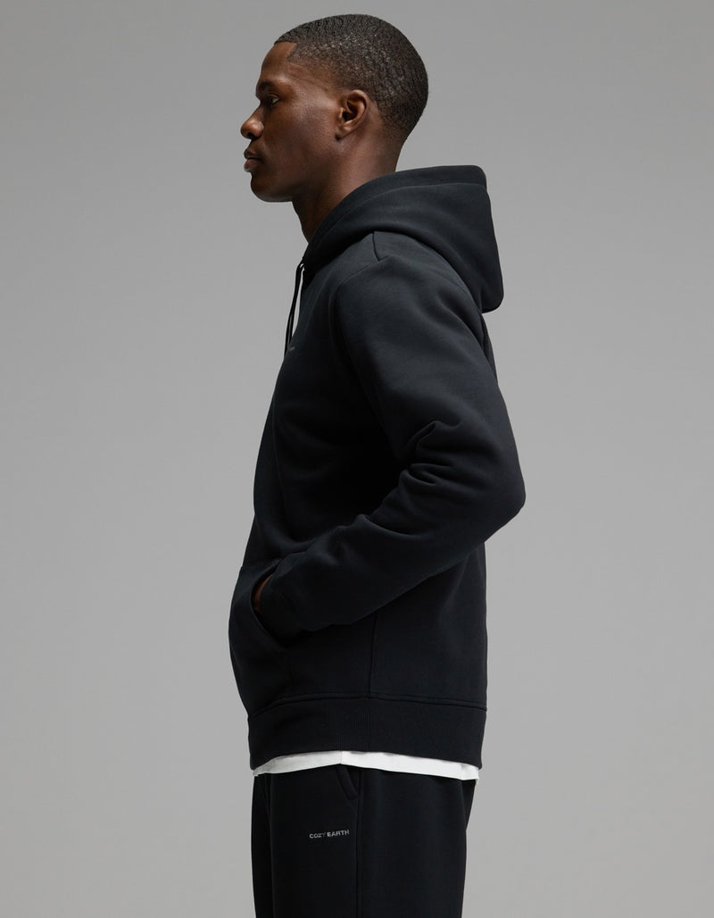 A man wearing the Cozy Earth Men’s Cityscape Hoodie & Sweatpant Set stands in profile against a plain gray background, his hands in the hoodie pocket as he looks forward.