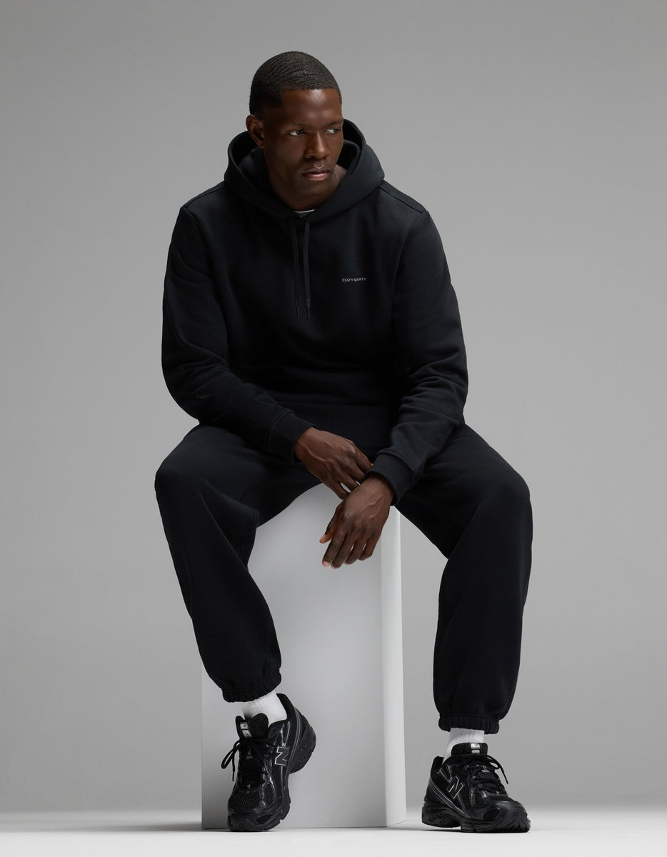 A man wearing the Cozy Earth Men’s Cityscape Hoodie & Sweatpant Set sits on a white block against a gray background, paired with black sneakers and white socks.