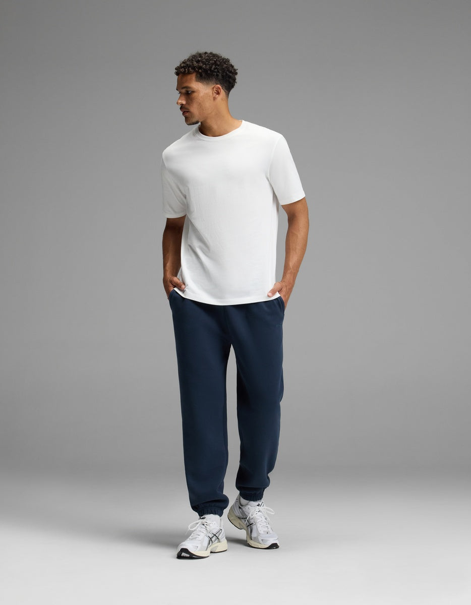 A man stands against a gray background, wearing a white t-shirt, white sneakers, and Cozy Earth’s Men’s Cityscape Sweatpant in navy blue, with his hands in his pockets and looking to the side.