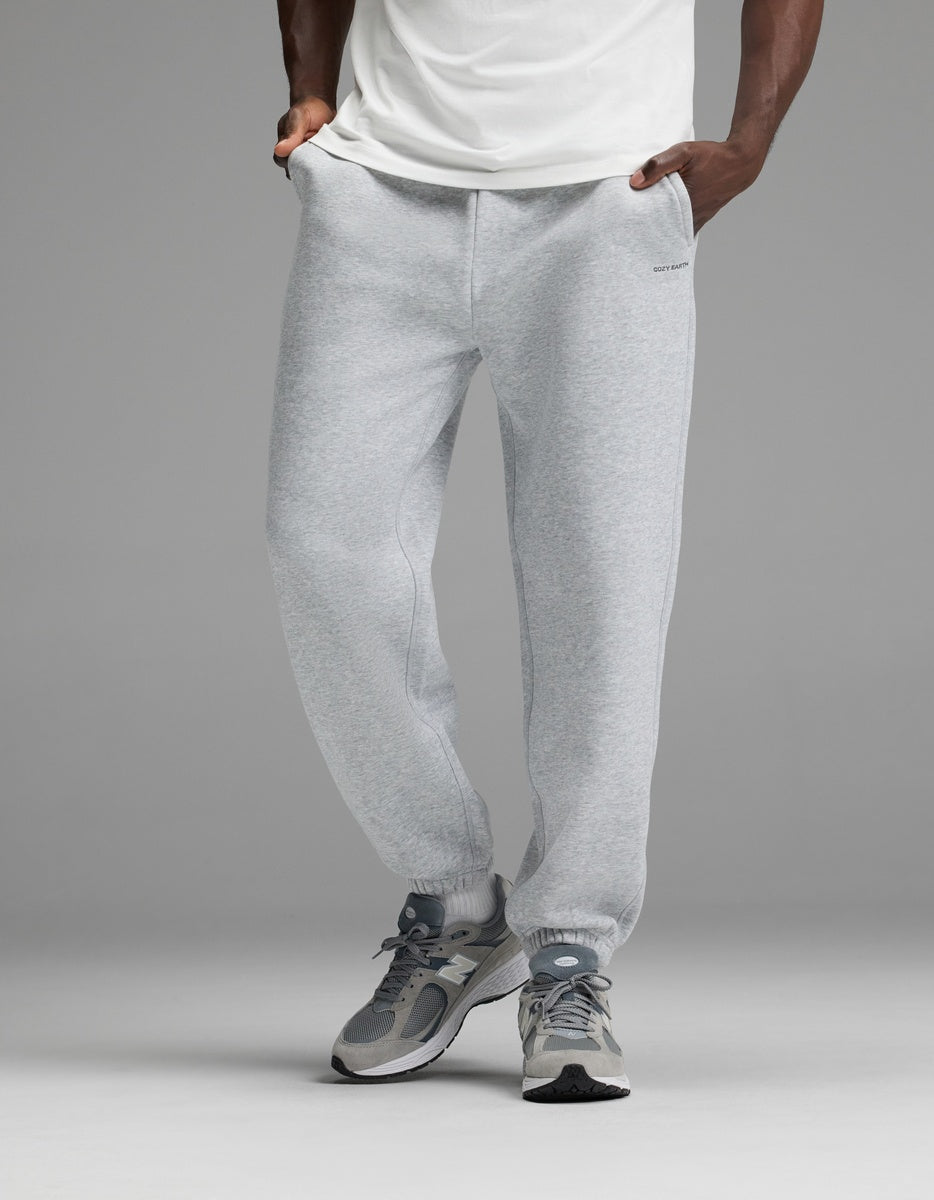 A person wearing a white shirt, Cozy Earth Men’s Cityscape Sweatpant in light gray, and gray sneakers stands with hands in pockets against a plain gray background.|Color:Heather Grey