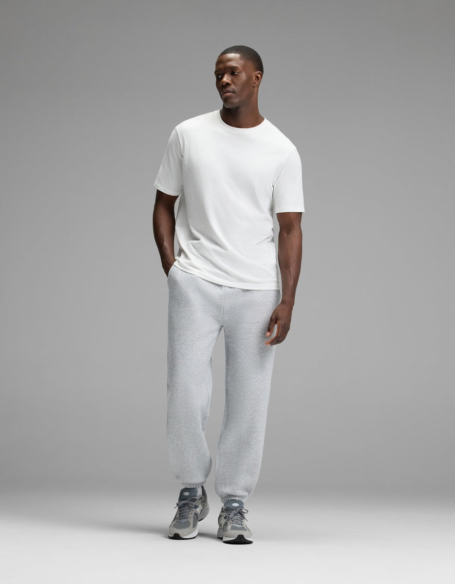 A man stands against a plain gray background, wearing a white t-shirt, Cozy Earth Men’s Cityscape Sweatpant in light gray, and gray sneakers. His hands are in his pockets, and he is looking slightly to the side with a neutral expression.|Color:Heather Grey