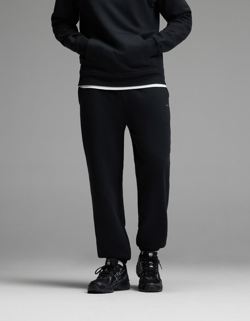 A person standing with hands in the pockets of Cozy Earth Men’s Cityscape Sweatpant in Jet Black, paired with a black sweatshirt, white shirt underneath, and black sneakers. The upper face is not visible.