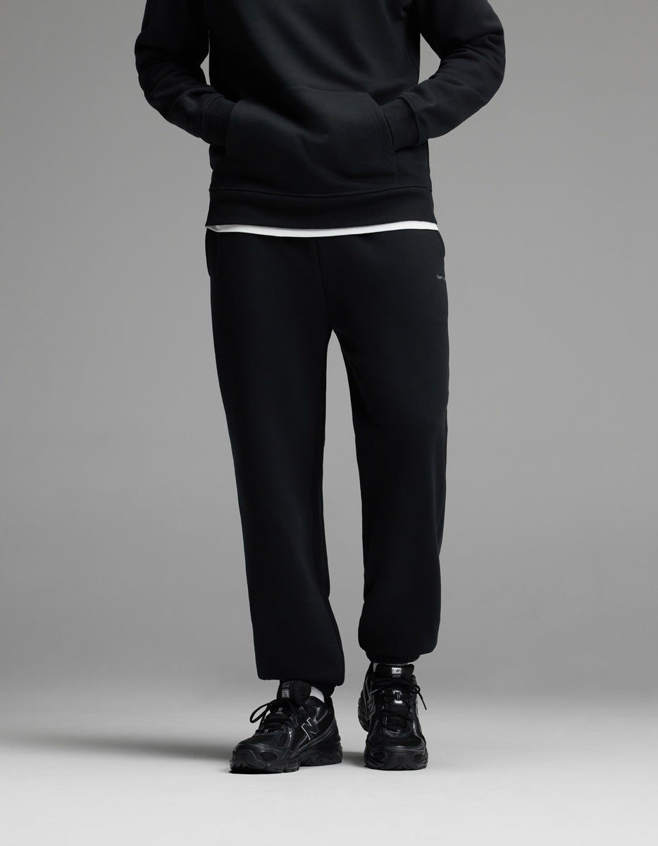A person standing with hands in the pockets of Cozy Earth Men’s Cityscape Sweatpant in Jet Black, paired with a black sweatshirt, white shirt underneath, and black sneakers. The upper face is not visible.|Color:Jet Black