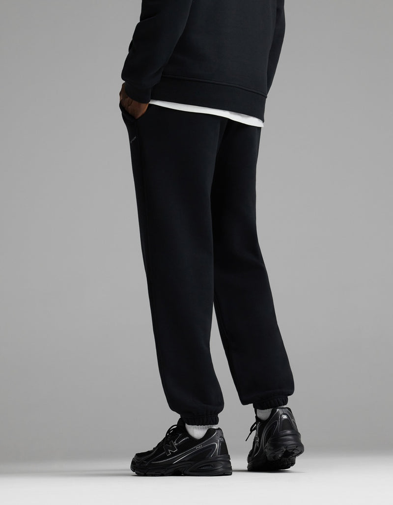 A person wearing Cozy Earth Men’s Cityscape Sweatpant in black stands facing away, with only their legs and lower torso visible, against a plain gray background.