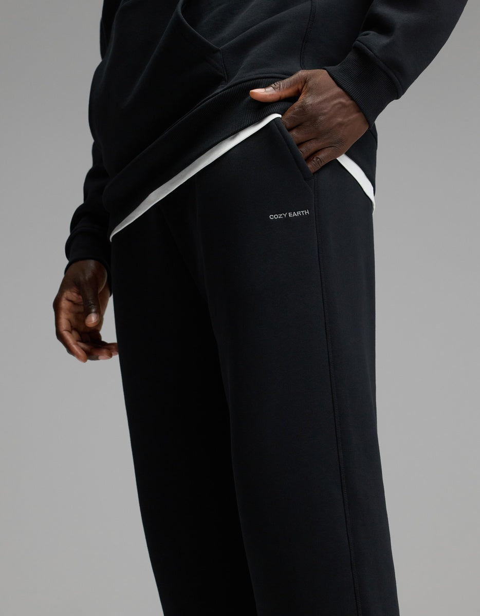 A person wearing the Cozy Earth Men’s Cityscape Sweatpant in black, paired with a matching black sweatshirt, stands with one hand in their pocket. The sweatpants feature “COZY EARTH” printed near the pocket, set against a plain gray background.