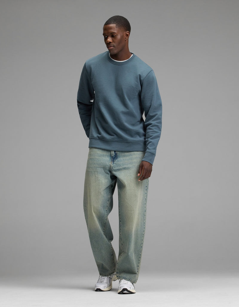 A person stands against a plain gray background wearing the Cozy Earth Men’s Dylan Crewneck, paired with loose light blue jeans and white sneakers, looking slightly downward with hands by their sides.