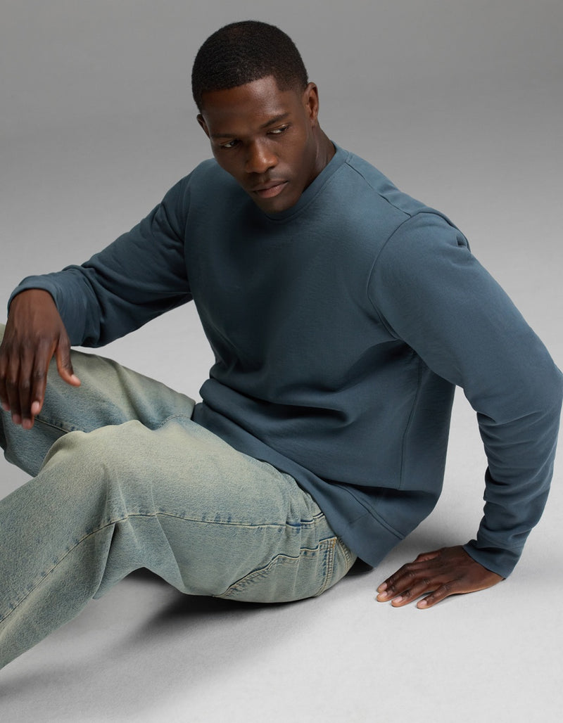 A man models the Cozy Earth Men’s Dylan Crewneck in blue, paired with light-wash jeans. He sits on the floor against a plain, neutral background, looking down with one hand on his knee and the other resting beside him.