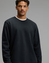 A man wearing the Men’s Dylan Crewneck in Jet Black by Cozy Earth over a white shirt stands against a plain gray background, looking toward the camera with a neutral expression.|Color:Jet Black